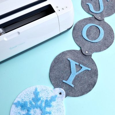 The new Cricut Maker cuts felt like butter! Learn how to make this DIY "Joy" Felt Winter Door Decoration in this no sew project that's perfect for a front door Christmas wreath substitute! How pretty are the sparkle snowflakes? #CricutMade #CricutHoliday #ChristmasCrafts #sponsored