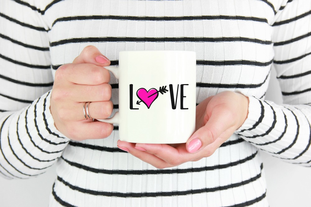 Woman holding a white mug with the word love on the front. The O is a heart with cupid's arrow through it. 