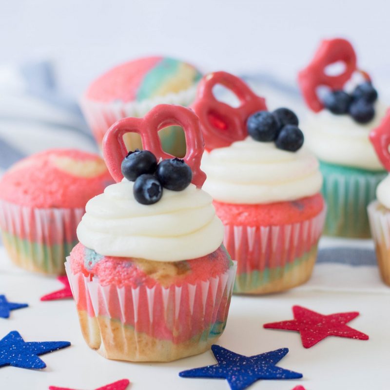 Celebrate the Fourth of July with this Patriotic Cupcakes Recipe! With red, white and blue swirls, buttercream frosting, hand dipped candy coated pretzels and blueberries, this is a fun Fourth of July Cupcake decorating idea! Great for other patriotic holidays too! Who doesn't love red, white and blue food ideas! #Cupcakes #FourthOfJuly #Patriotic #Baking #CupcakeDecorating #Recipe #CupcakeRecipe #RedWhiteAndBlue #PatrioticFood