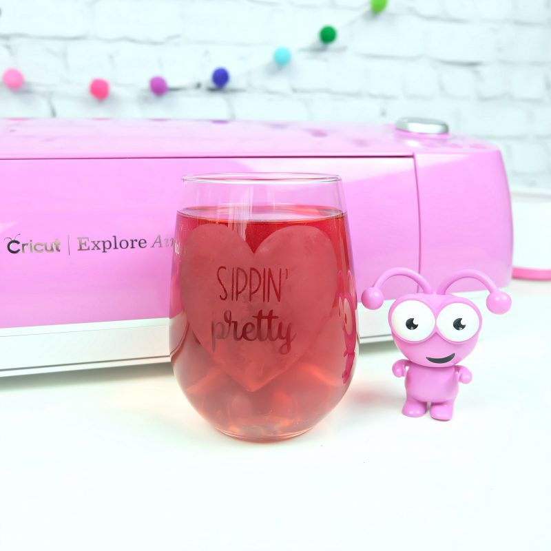 A pink Cricut Cutie stands in front of a fuchsia Cricut Explore Air 2 and a white brick wall with a felt pom pom garland. He stands next to a wine tumbler filled with pink liquid that has Sippin' Pretty etched in it. Project and cut file from the Book The Unofficial Book of Cricut Crafts