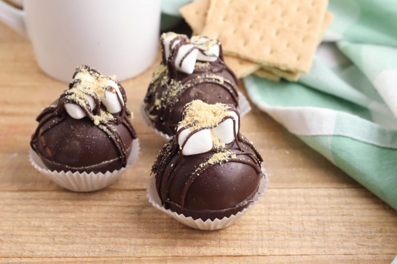 S'mores hot chocolate bombs laying on a wood board with graham crackers and a mug.