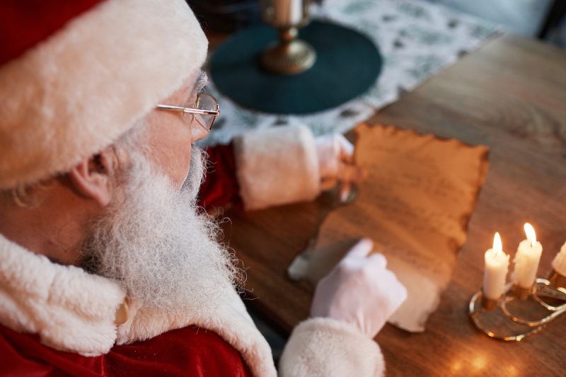 Over shoulder view of Santa Claus in glasses sitting at table and reading letter under candlelit