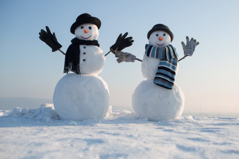 Two snowmen sitting in the snow dressed in hats, gloves and scarves. 