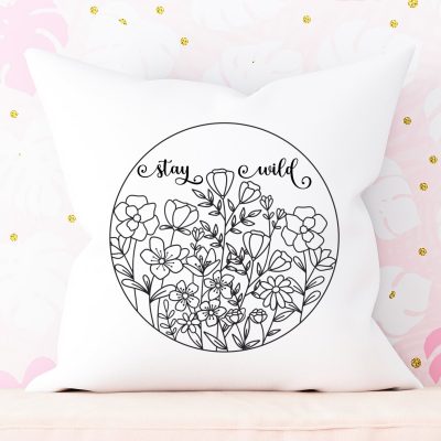 White throw pillow leaned against wall with pink and white monstera leaf wallpaper. Throw pillow has a design with wildflowers in a circle with the words Stay Wild above.