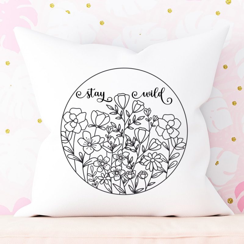 White throw pillow leaned against wall with pink and white monstera leaf wallpaper. Throw pillow has a design with wildflowers in a circle with the words Stay Wild above. 