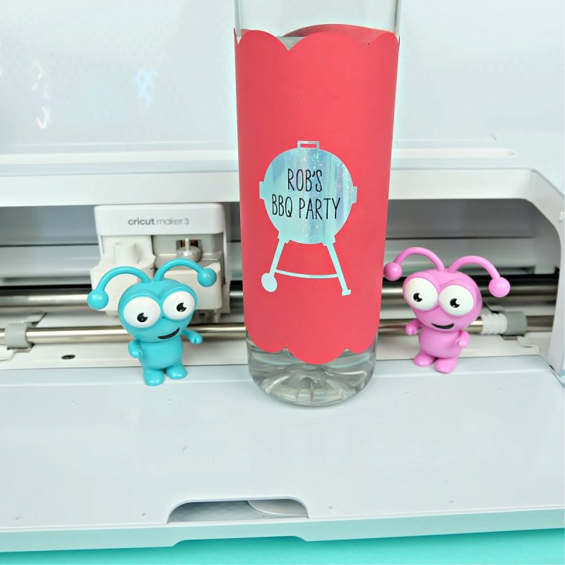Water bottle with DIY wrap sitting in front of a Cricut Maker 3 with two Cricut cuties