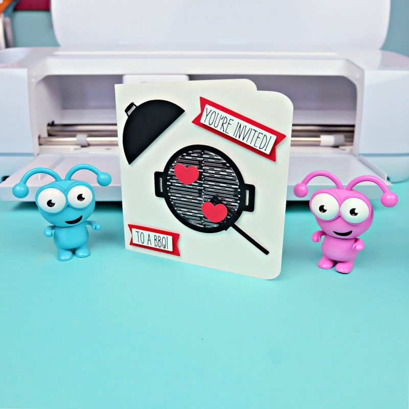Invitation that reads "You are invited to a BBQ" sitting in front of a Cricut cutting machine with two Cricut cuties.