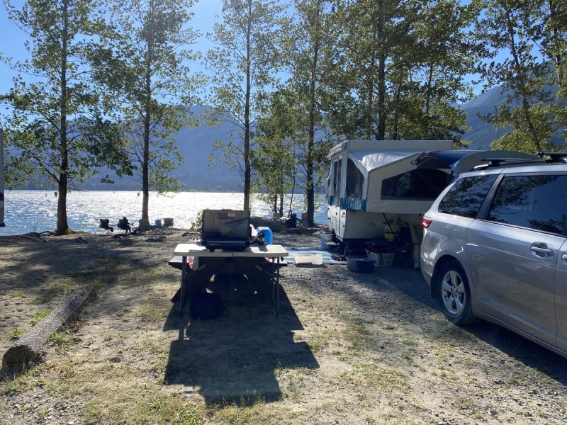 Sparkling blue lake with trees, foothills and a camping spot with a tent trailer set up. 