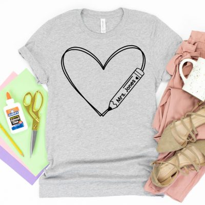 An image of a grey T-shirt with a heart and pencil design on the front. The shirt is surrounded by various school supplies, including paper, glue, scissors and a coffee mug.
