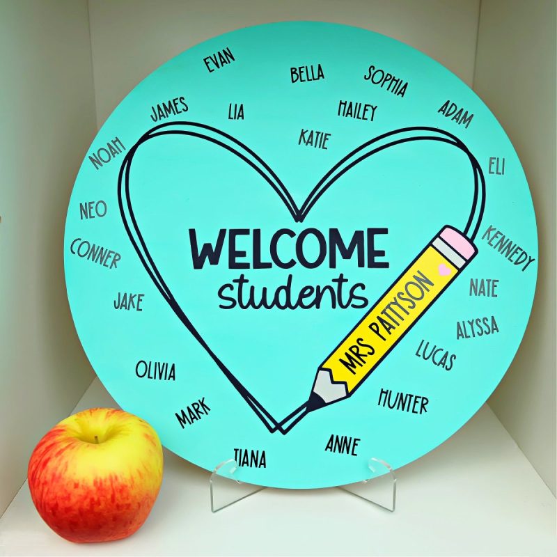 DIY Classroom Sign sitting on a stand on a shelf with an apple next to it. Sign is blue, says welcome students and has each of the students names as well as the teacher's name.