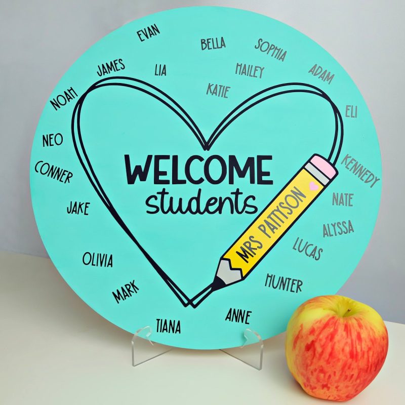 DIY Classroom Sign sitting on a stand on a table with an apple next to it. Sign is blue, says welcome students and has each of the students names as well as the teacher's name.