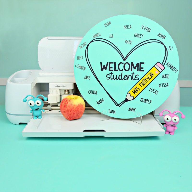 DIY Classroom Sign sitting on a stand on a Cricut Maker 3 with an apple next to it. Sign is blue, says welcome students and has each of the students names as well as the teacher's name.