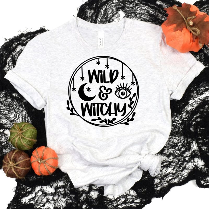 Heather grey t-shirt laying on black fabric surrounded by pumpkins. A halloween design that says Wild & Witchy made from black HTV is on the front. 