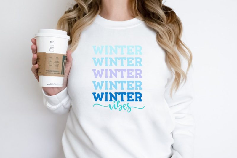 Woman in white sweatshirt with coffee cup. Sweatshirt has Winter Vibes in pretty winter shades of blue on the front. 