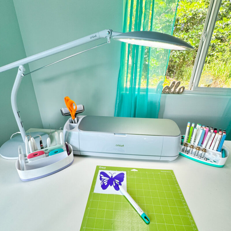 Cricut Bright 360 Lamp on a table with a Cricut and Cricut Cutting Mat with a weeded piece of vinyl on top.