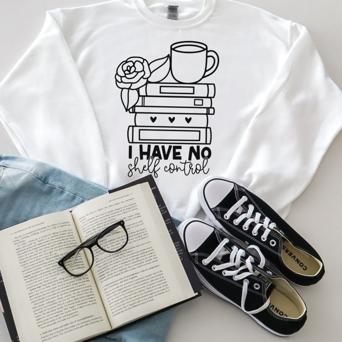 Sweatshirt that reads I Have No Shelf Control with a stack of books with a mug and flower on top. Image created by Hello Creative Family