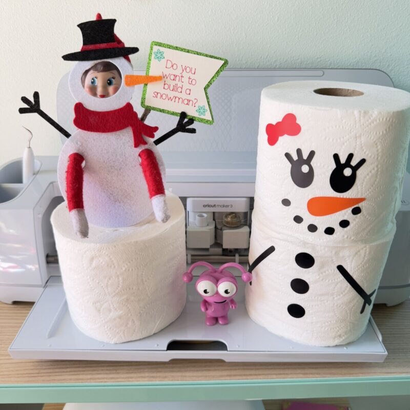 Elf on the Shelf dressed in a handmade felt snowman costume, standing next to toilet paper rolls transformed into snowmen with Cricut vinyl stickers with a Cricut Maker in the background.