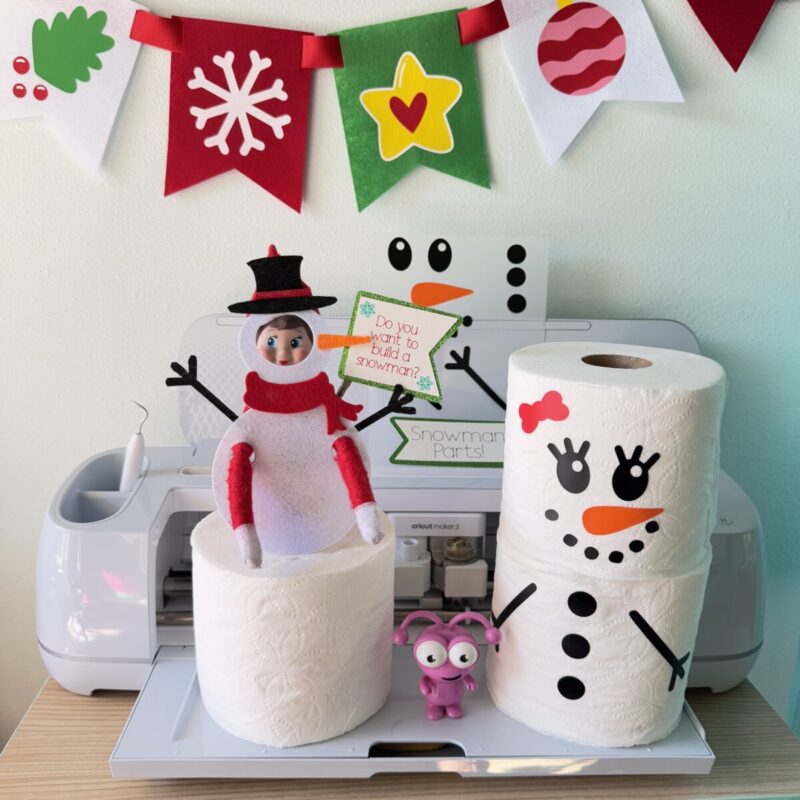Elf on the Shelf dressed in a handmade felt snowman costume, standing next to toilet paper rolls transformed into snowmen with Cricut vinyl stickers with a Cricut Maker in the background.