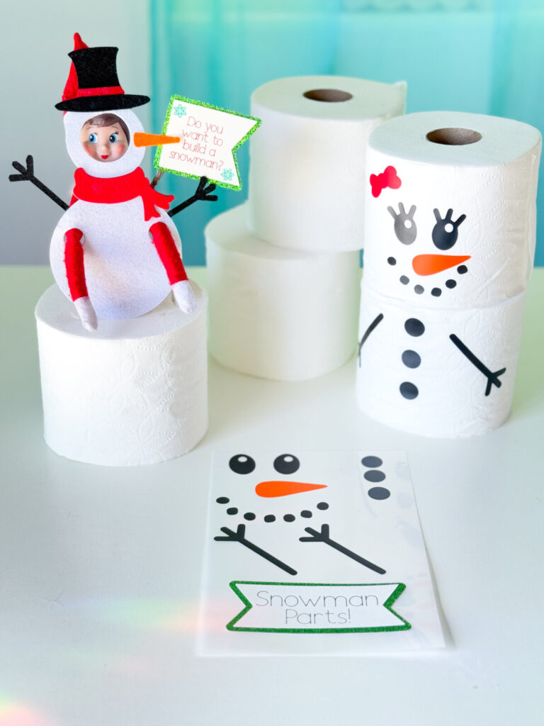 DIY Elf on the Shelf snowman scene featuring an elf in a snowman costume and snowmen made from toilet paper rolls with vinyl decals.