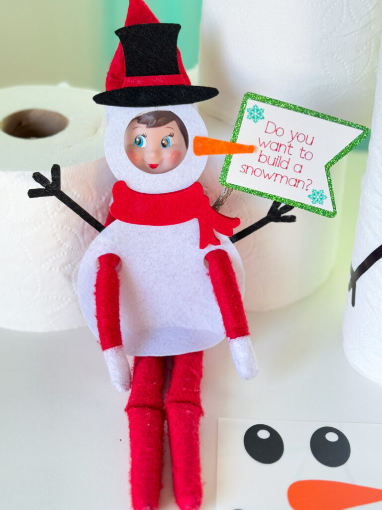 Close-up of an Elf on the Shelf wearing a snowman outfit made from felt, with toilet paper snowmen decorated with carrot nose and button decals.