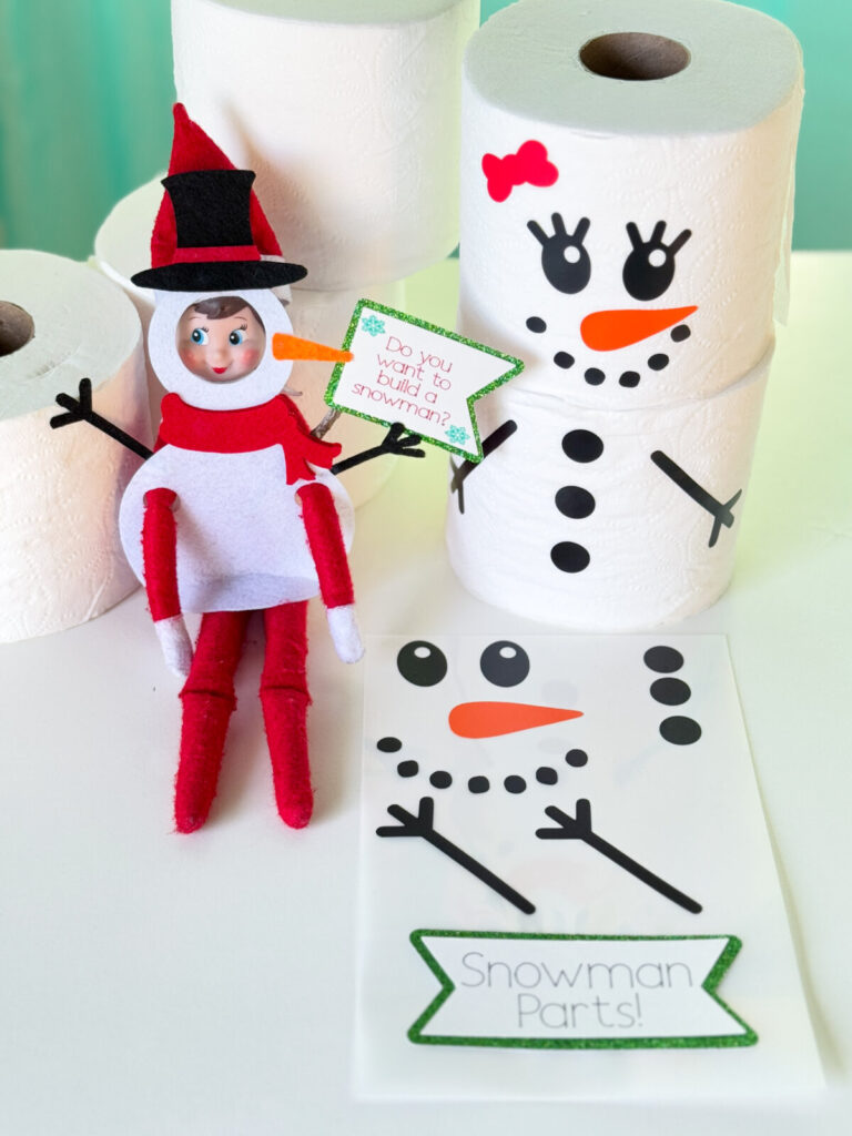 Close-up of an Elf on the Shelf wearing a snowman outfit made from felt, with toilet paper snowmen decorated with carrot nose and button decals.