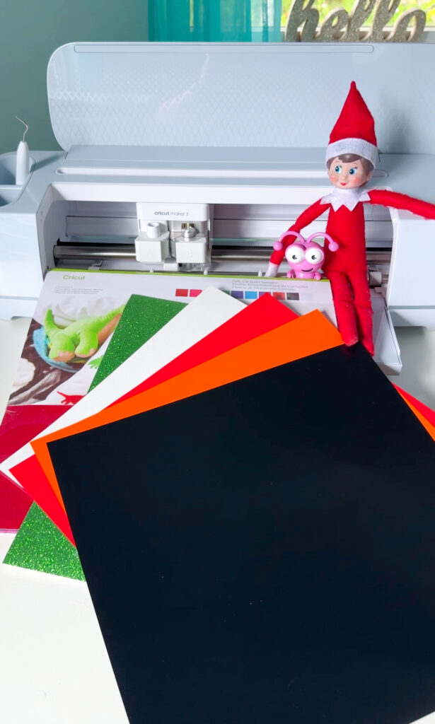 Elf on the shelf sitting on a Cricut Maker 3 with Cricut craft supplies