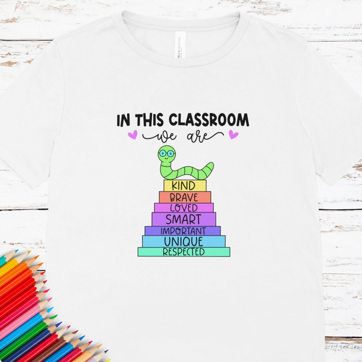 Teacher t-shirt that reads "In This Classroom We Are Kind, Brave, Loved, Smart, Important, Unique, Respected." The words of affirmation are on a stack of books with a bookworm posed on top.