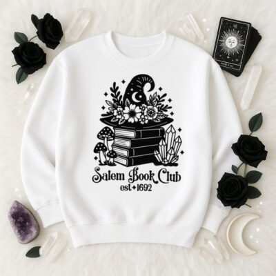 White sweatshirt with a black &ldquo;Salem Book Club est. 1692&rdquo; SVG design showing a witch hat decorated with flowers on a stack of books, surrounded by mushrooms, crystals, and stars. Displayed on a white faux fur background with tarot cards, black roses, crystals, amethyst, and a crescent moon.