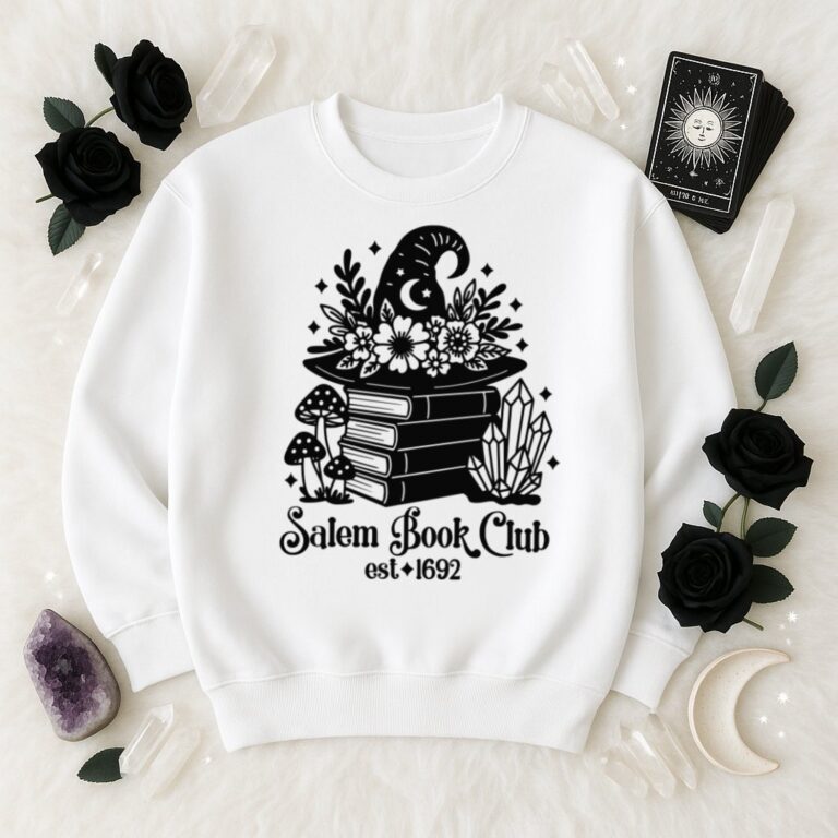 White sweatshirt with a black “Salem Book Club est. 1692” SVG design showing a witch hat decorated with flowers on a stack of books, surrounded by mushrooms, crystals, and stars. Displayed on a white faux fur background with tarot cards, black roses, crystals, amethyst, and a crescent moon.