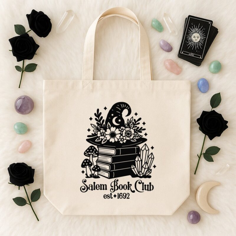 Canvas tote bag with a black “Salem Book Club est. 1692” SVG design featuring a witch hat adorned with flowers, stacked books, mushrooms, crystals, and stars. Styled on a white faux fur background with tarot cards, gemstones, black roses, and a crescent moon prop.