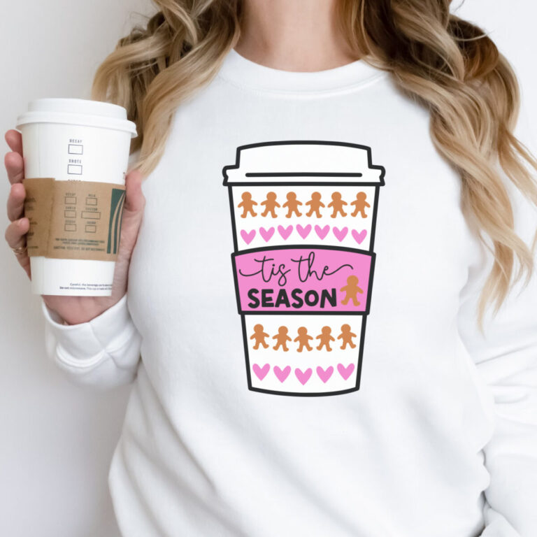 A woman wearing a white sweatshirt featuring a festive “Tis The Season” coffee cup design with rows of gingerbread men and pink hearts, holding a to-go coffee cup in her hand. The image conveys cozy Christmas vibes and is perfect for showcasing a holiday SVG design for Cricut projects.