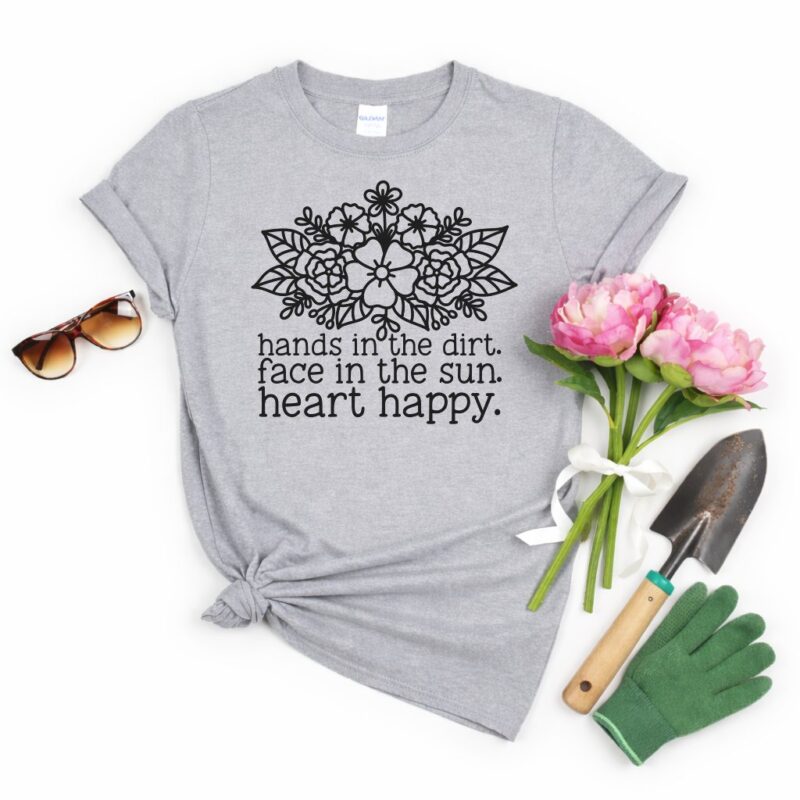 Flat lay of a heather grey t-shirt featuring a black floral design and the quote “Hands In The Dirt. Face In The Sun. Heart Happy.” styled with pink peonies, a garden trowel, green gardening gloves, and sunglasses on a white background.