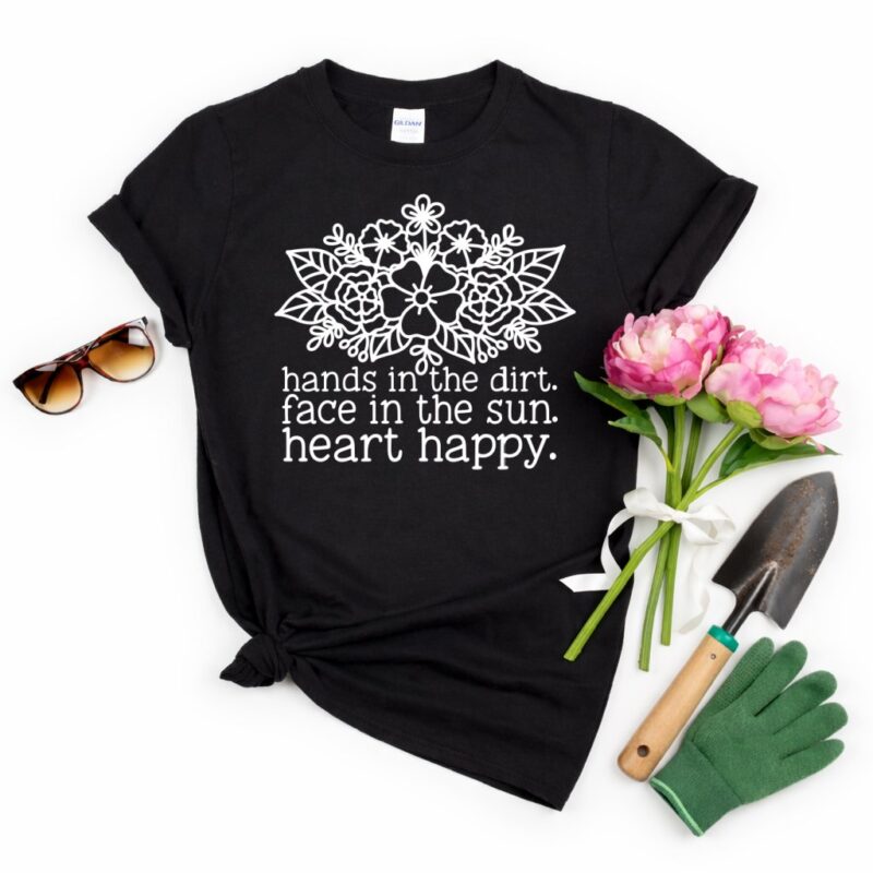Flat lay of a black t-shirt featuring a white floral design and the quote “Hands In The Dirt. Face In The Sun. Heart Happy.” styled with pink peonies, a garden trowel, green gardening gloves, and sunglasses on a white background.