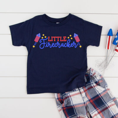 Navy toddler t-shirt featuring a colorful Little Firecracker SVG design with firework rockets, script lettering, and gold stars in red, white, and blue, styled with plaid shorts and patriotic decorations on a white shiplap background.