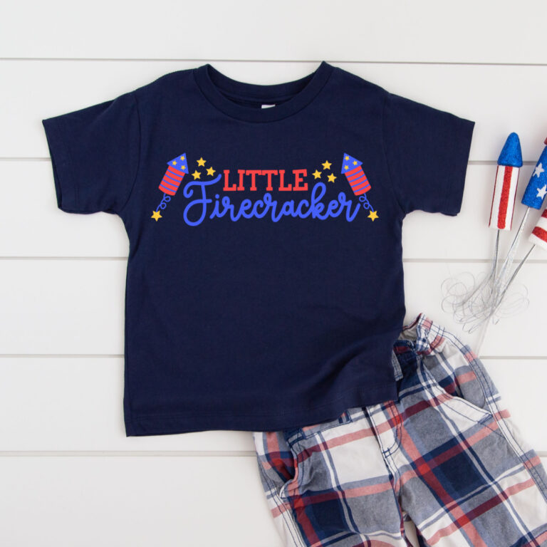 Navy toddler t-shirt featuring a colorful Little Firecracker SVG design with firework rockets, script lettering, and gold stars in red, white, and blue, styled with plaid shorts and patriotic decorations on a white shiplap background.