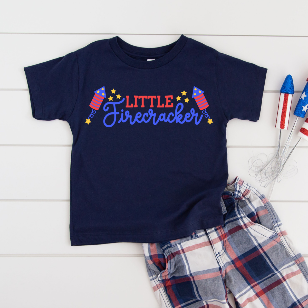 Navy toddler t-shirt featuring a colorful Little Firecracker SVG design with firework rockets, script lettering, and gold stars in red, white, and blue, styled with plaid shorts and patriotic decorations on a white shiplap background.